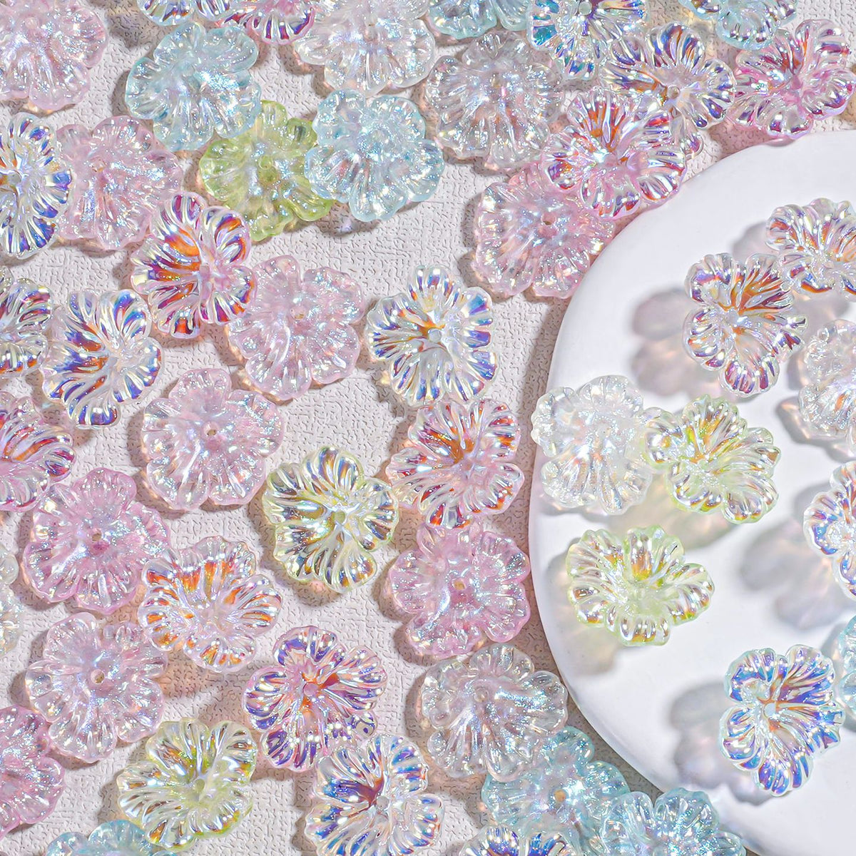 Iridescent resin hibiscus flower beads in pastel pink, blue and green scattered on white fabric