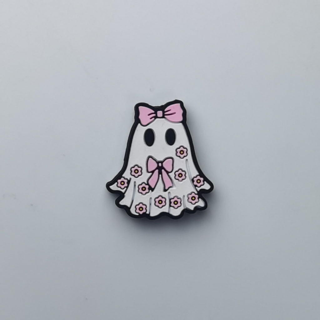 A cute ghost-shaped accessory, likely an enamel pin or charm, featuring a white body decorated with pink bows and small pink flowers, outlined in black, on a light gray background.