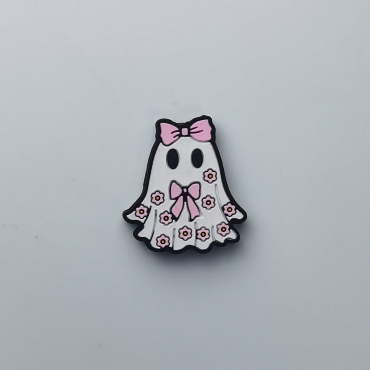 A cute ghost-shaped accessory, likely an enamel pin or charm, featuring a white body decorated with pink bows and small pink flowers, outlined in black, on a light gray background.