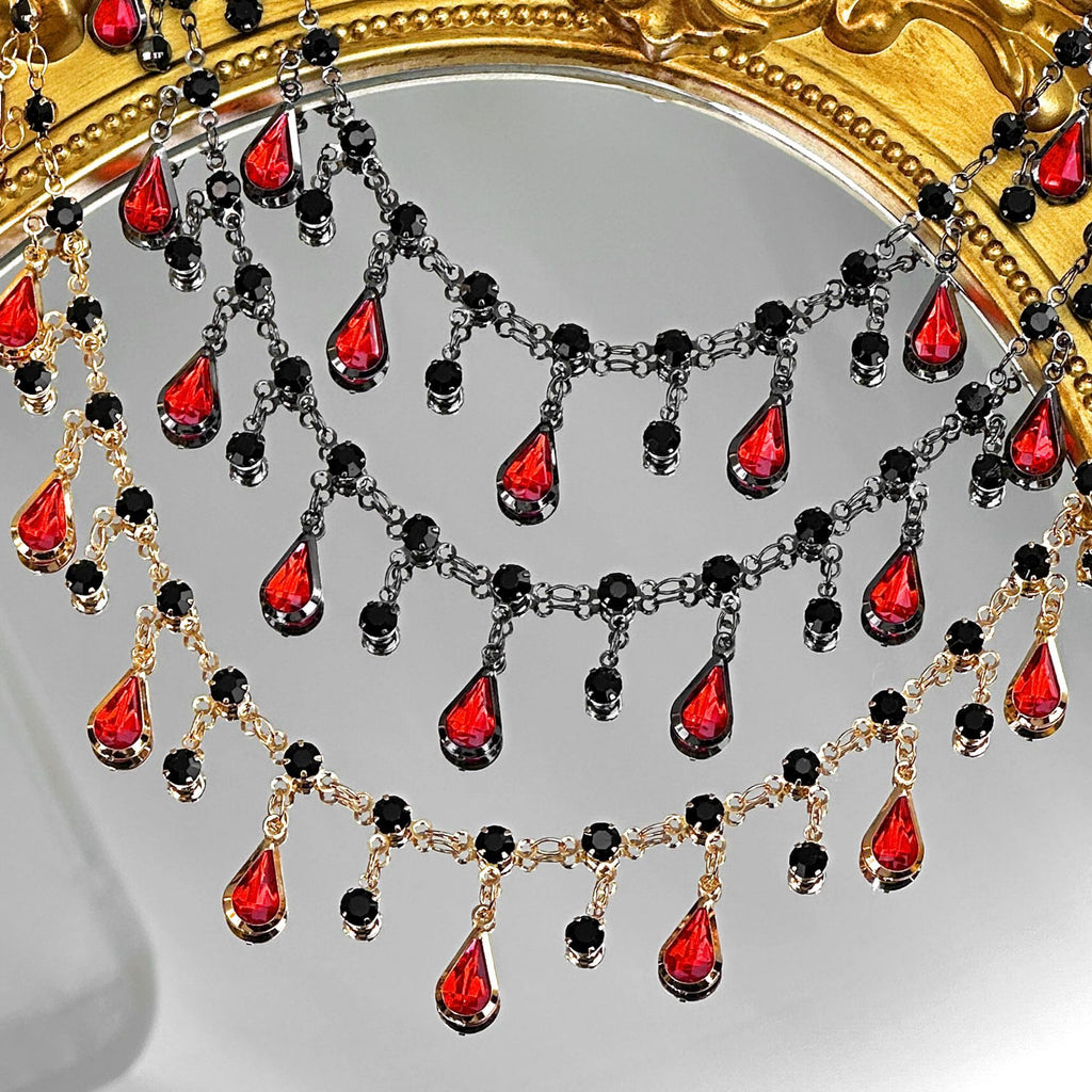 A close-up of an ornate gold mirror frame decorated with layered chains of red teardrop crystals and black round beads, hanging against the reflective mirror surface.