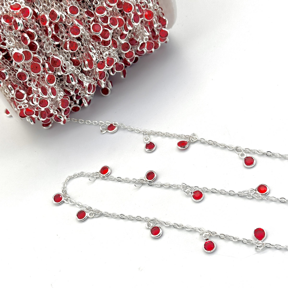A close-up of a silver chain embellished with small red circular studs, shown with a spool of matching chain in the upper left and several loose sections laid out on a white background.