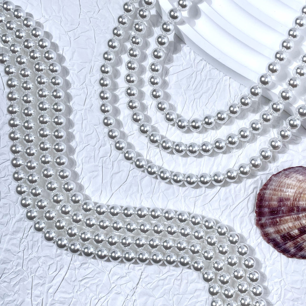 Silver-white glass pearl bead strands in S-curves on white textured surface with seashell prop