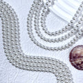 Silver-white glass pearl bead strands in S-curves on white textured surface with seashell prop