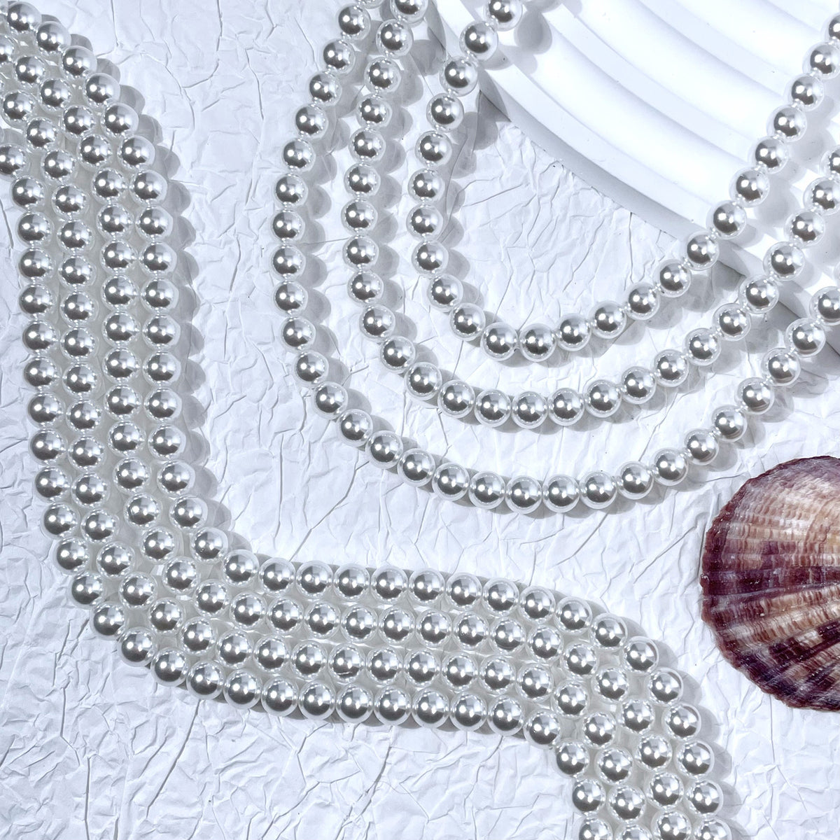 Silver-white glass pearl bead strands in S-curves on white textured surface with seashell prop