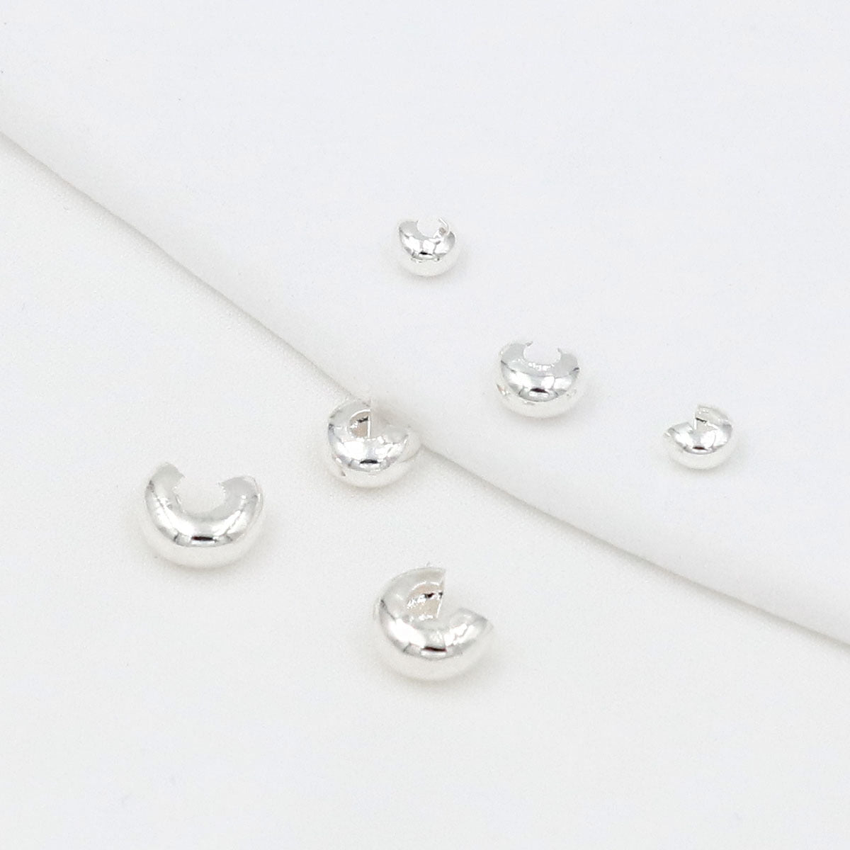 Six small polished silver C-shaped crimp bead covers scattered on a white background with a soft fold