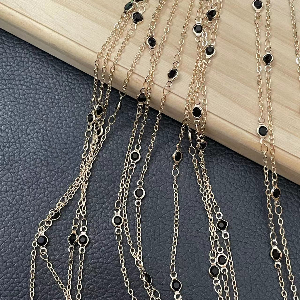 A close-up of multiple gold-tone chain necklaces draped over a dark textured surface, with small black oval and marquise-shaped stones set along the chains and a light wood block behind them.
