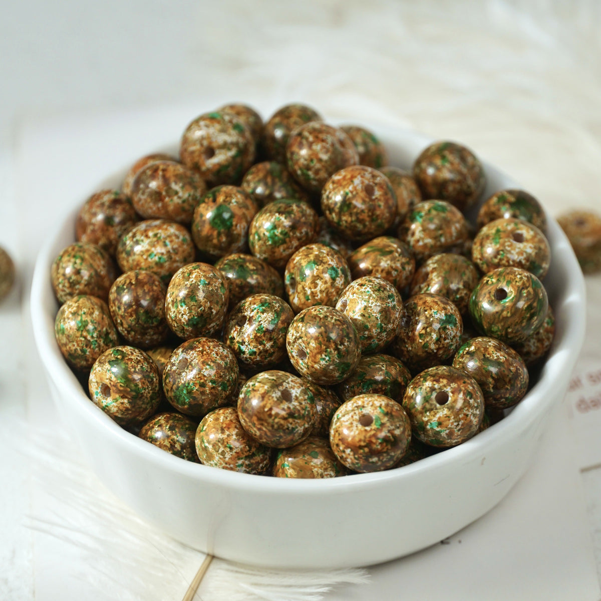 White porcelain bowl of round glossy brown beads speckled with gold and green on a soft light background