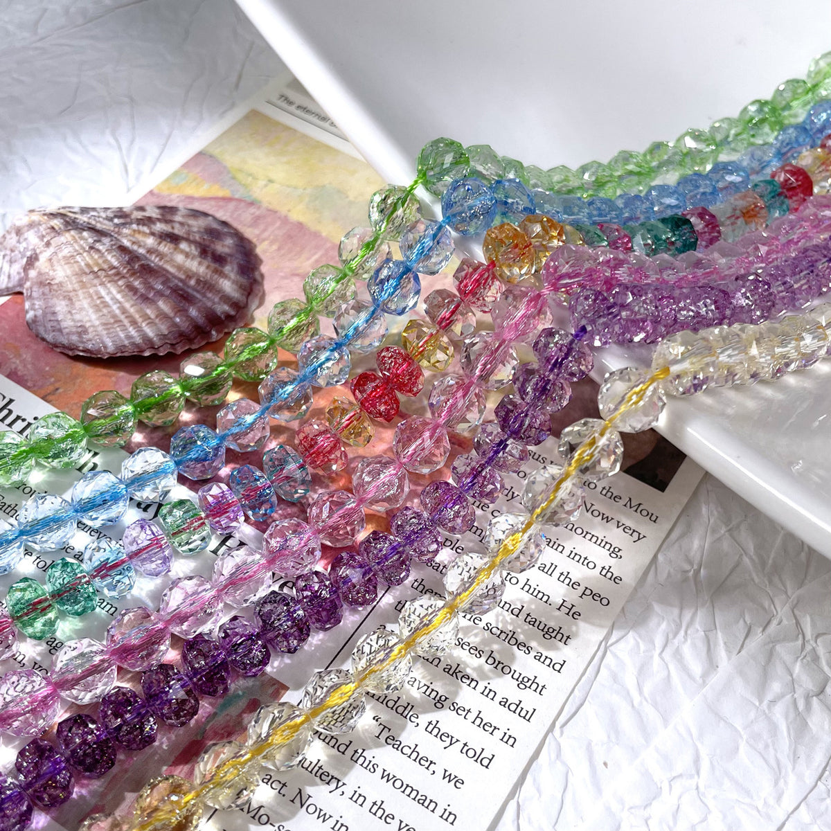 Multiple strands of faceted floral-crackle glass beads in assorted colors on an open book with a seashell nearby