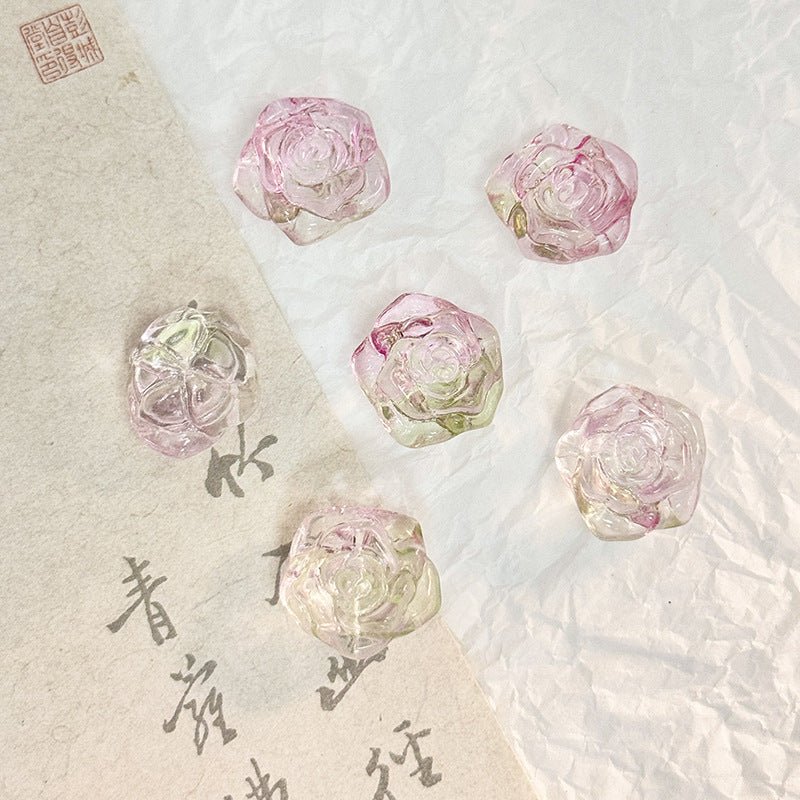 Five translucent pink rose-shaped glass beads on a textured white surface with a beige calligraphy paper strip