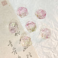 Five translucent pink rose-shaped glass beads on a textured white surface with a beige calligraphy paper strip