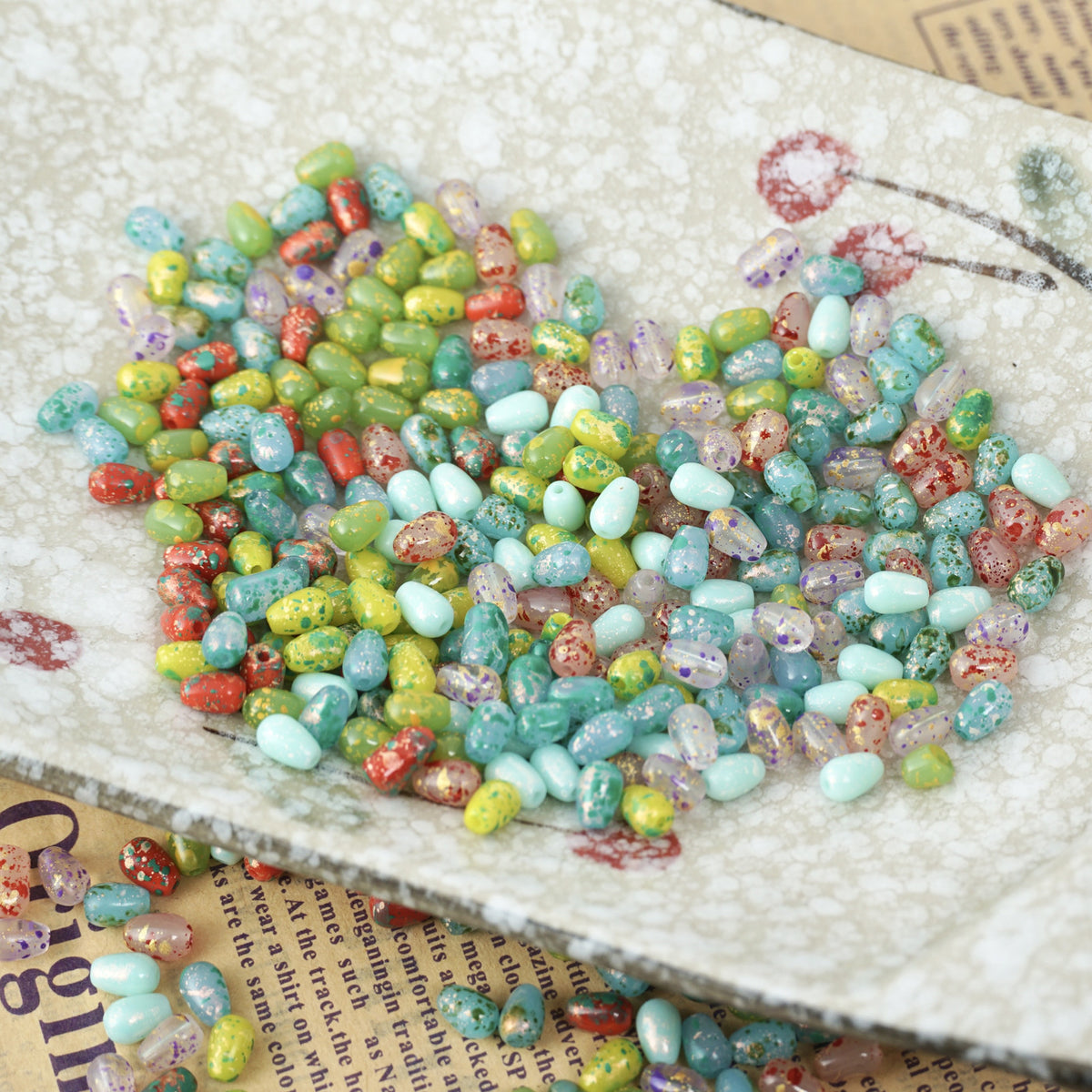 Colorful speckled glass teardrop beads on a white heart-shaped dish over a book page