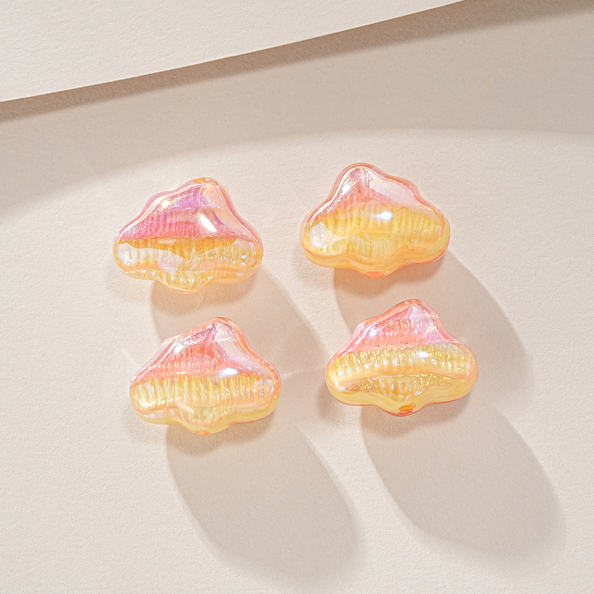 Four glossy iridescent cloud-shaped resin beads in pink, orange, and yellow on a beige surface.