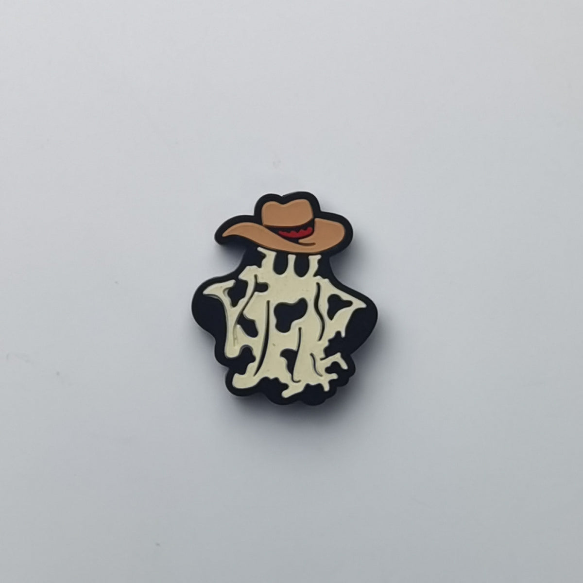 A small cartoon-style pin or charm featuring a tan cowboy hat with a red mouth and a cream-and-black cow-print body, centered on a plain light gray background.