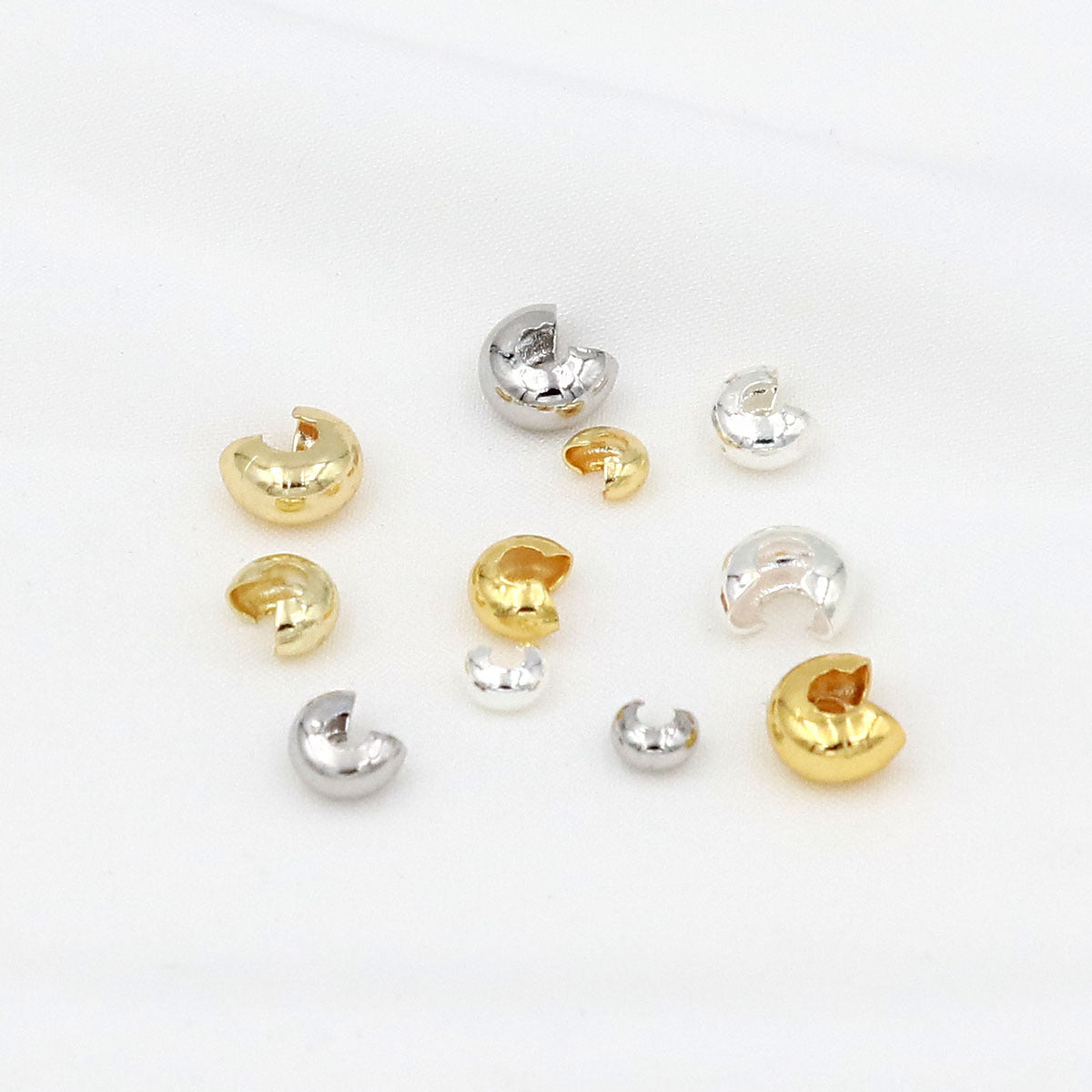 Small C-shaped crimp bead covers in polished gold and silver finishes arranged on a white background