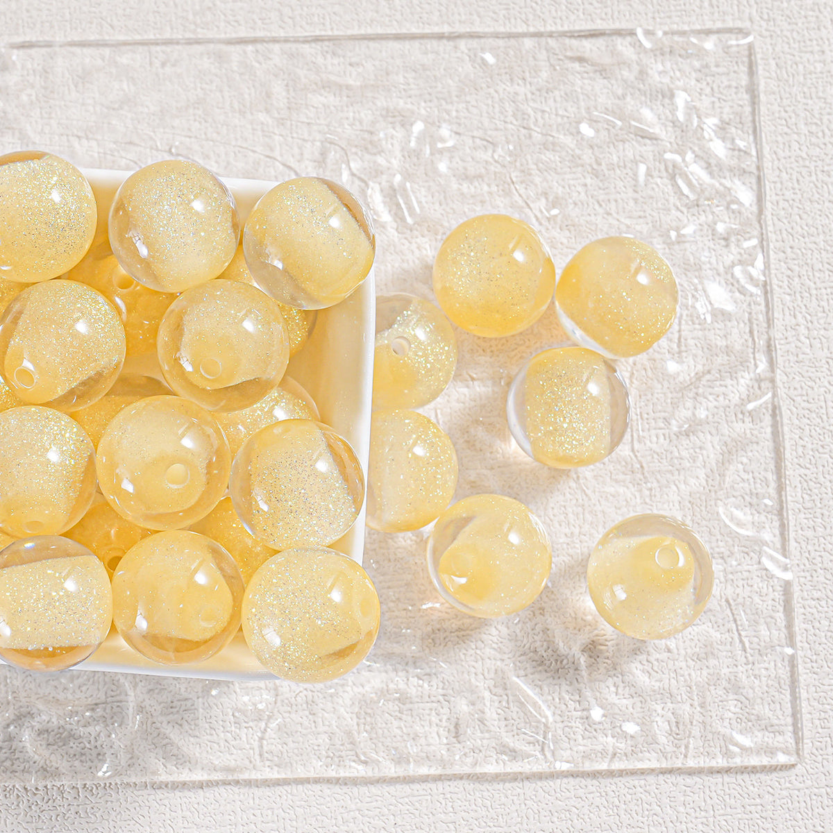 Yellow gold glitter round beads in a white tray and scattered on textured white surface