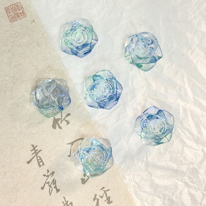 Five faceted translucent light blue and green rose-shaped glass beads on white and beige paper