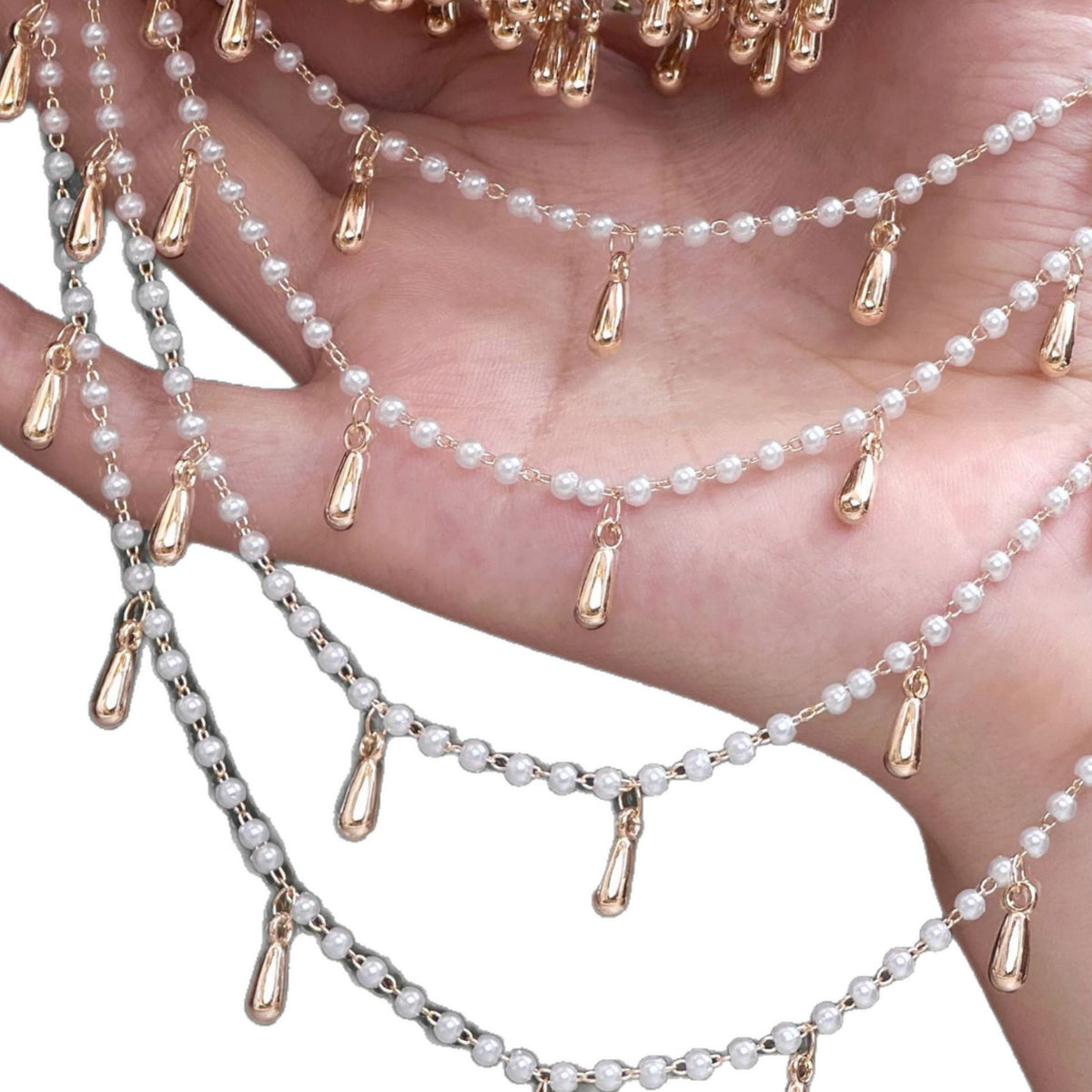 Hand holding layered faux pearl chains with rose gold teardrop charms on a white background