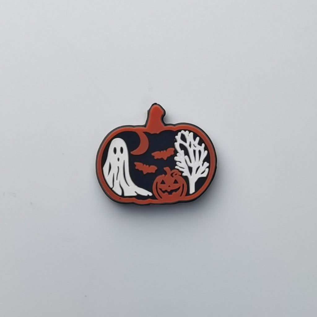 A Halloween-themed enamel pin shaped like a pumpkin, featuring a white ghost, jack-o'-lantern, bats, a tree silhouette, and a crescent moon on a black background with an orange border, shown on a light gray surface.