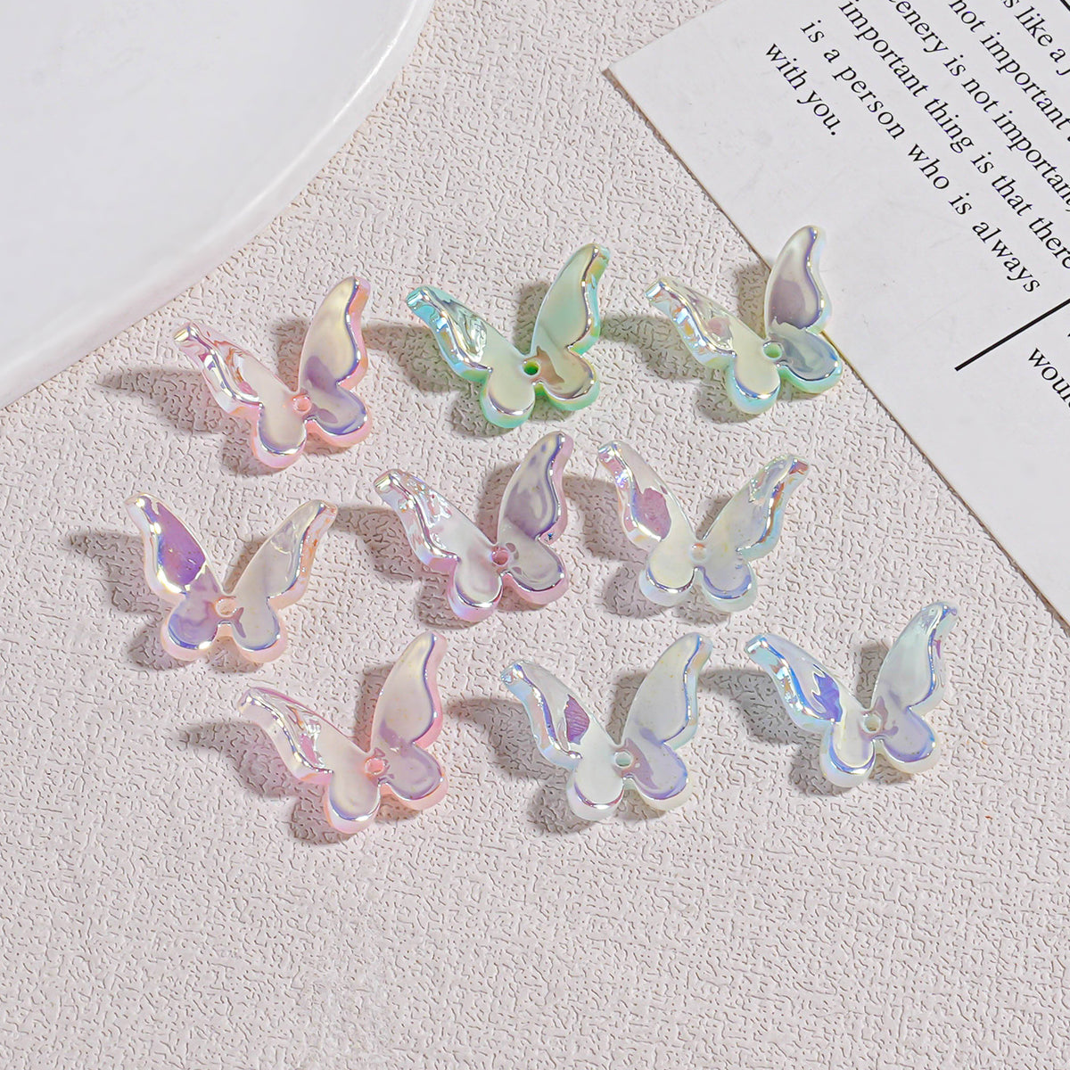 Twelve iridescent butterfly acrylic beads on textured beige surface with plate and paper