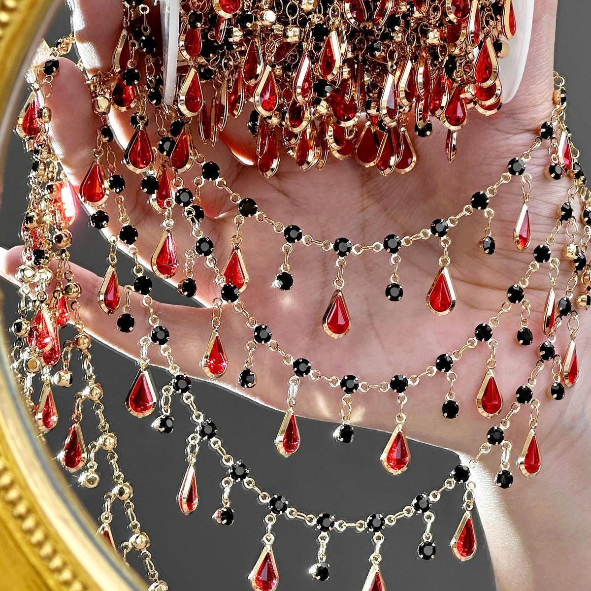 A close-up of a hand holding a gold-toned jewelry stand or display, with multiple draped chain necklaces featuring red teardrop gemstones and small black round stones. The background is dark gray with a blurred gold mirror edge visible on the left.
