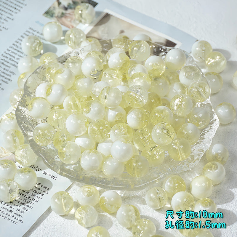 Pale yellow and white ice-crackle glass beads piled in a clear glass dish on a white surface with paper props