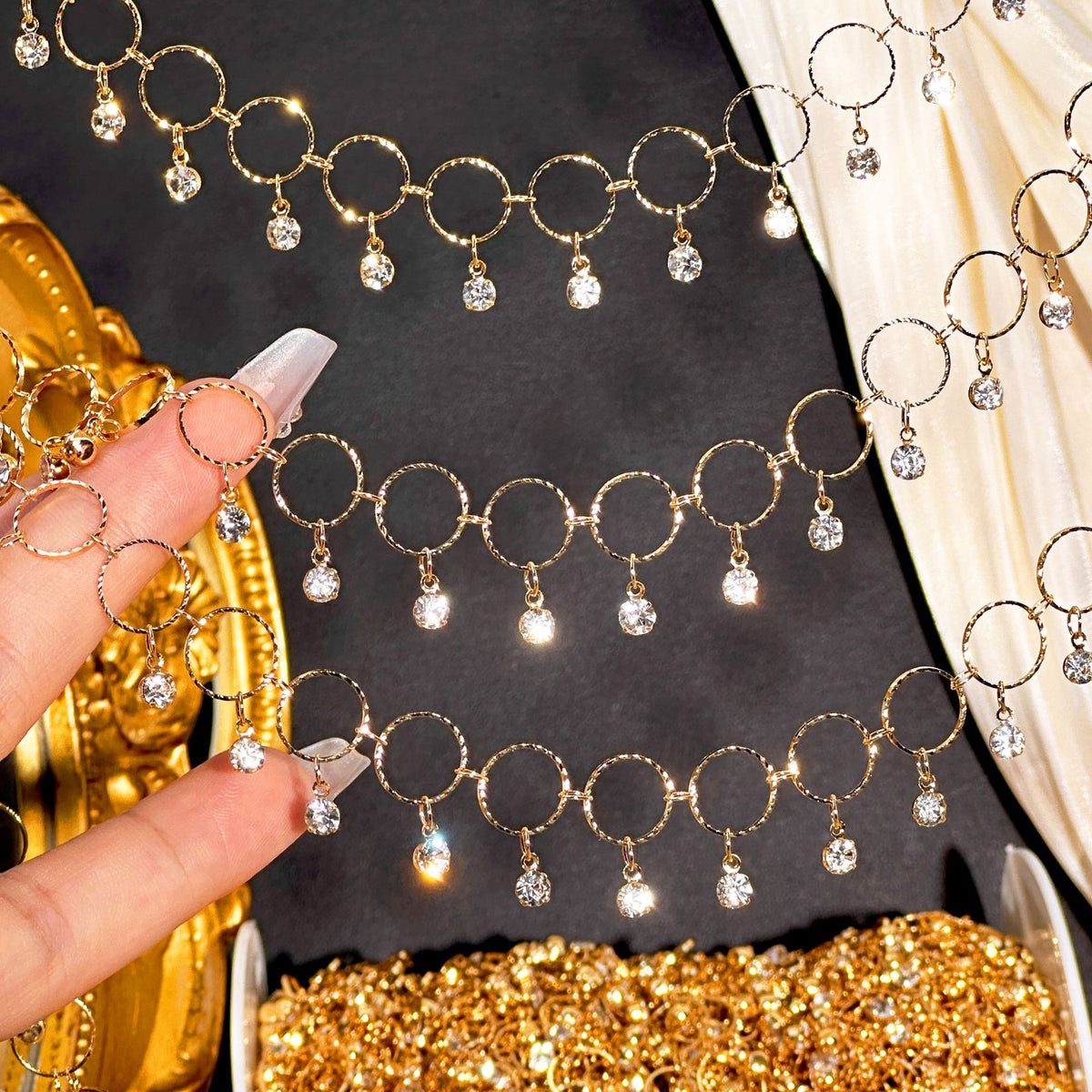 Gold chain necklace with circular links and dangling clear crystal charms, shown draped across a dark surface with a hand holding a section and a gold jewelry box visible.