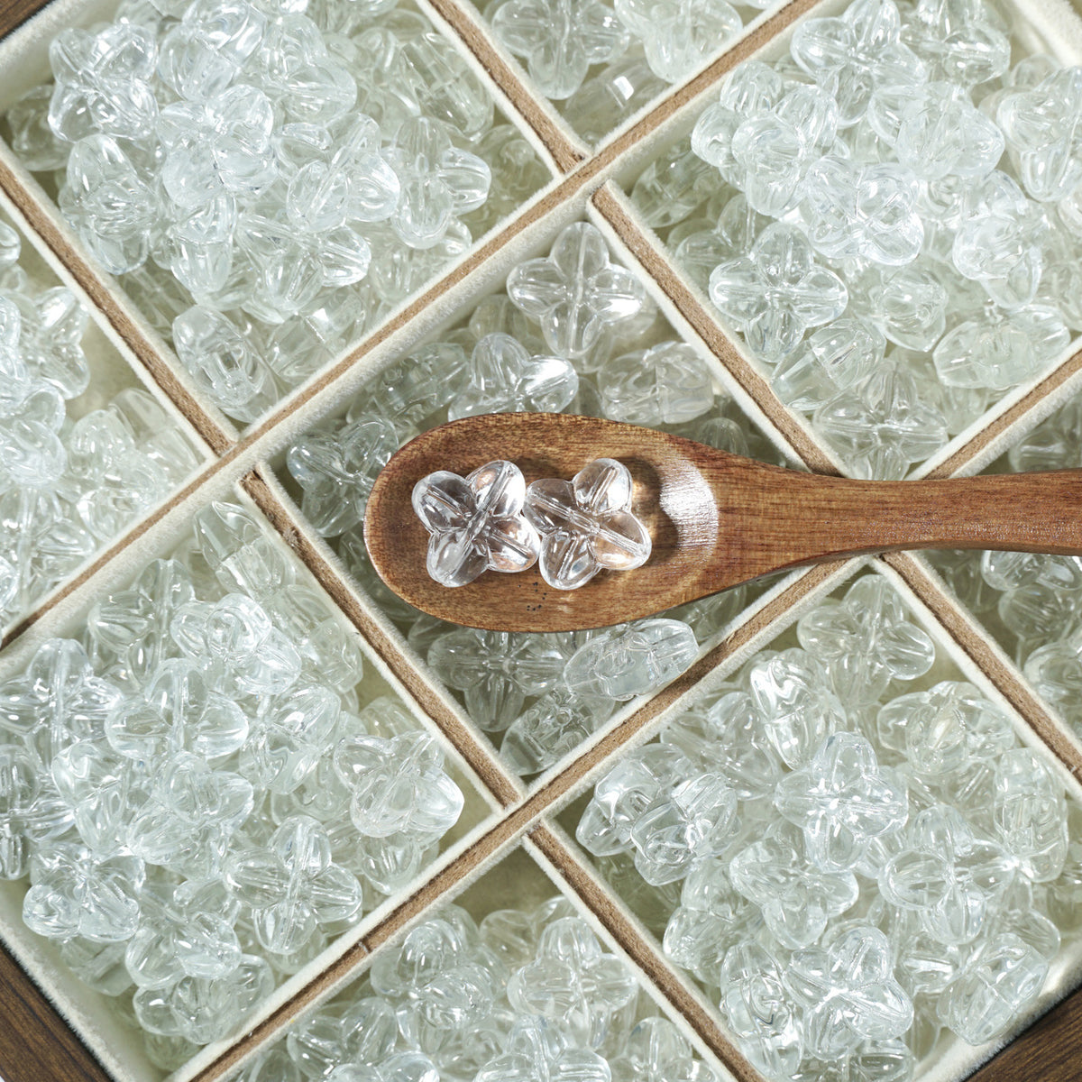 Clear flower-shaped glass beads in a beige compartment box, three beads on a small wooden spoon