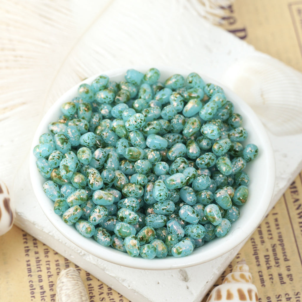 Speckled turquoise-blue glass teardrop beads in a white ceramic bowl on vintage book pages with shells nearby.