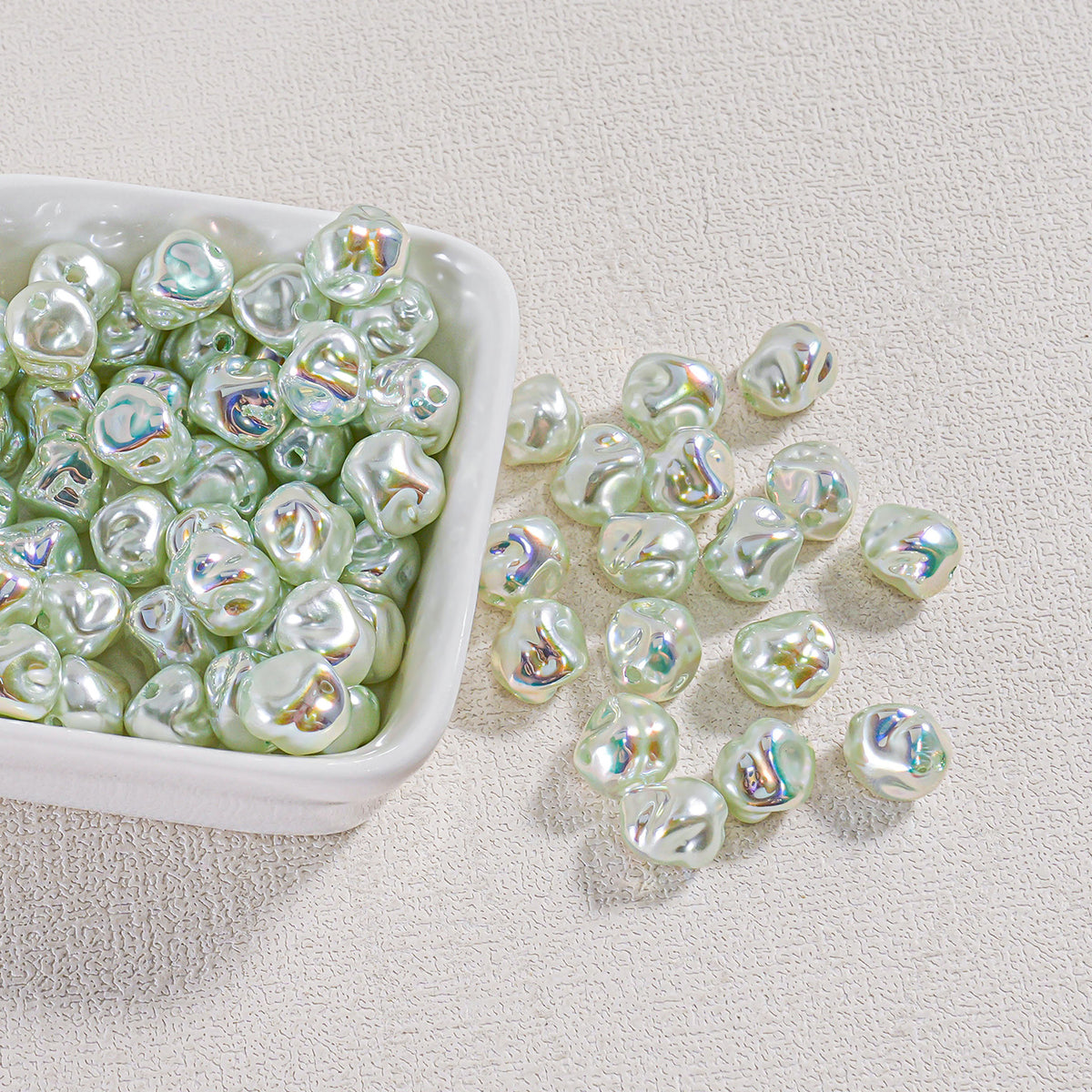 Iridescent green baroque faux pearl beads in a white square dish with several scattered on a textured surface
