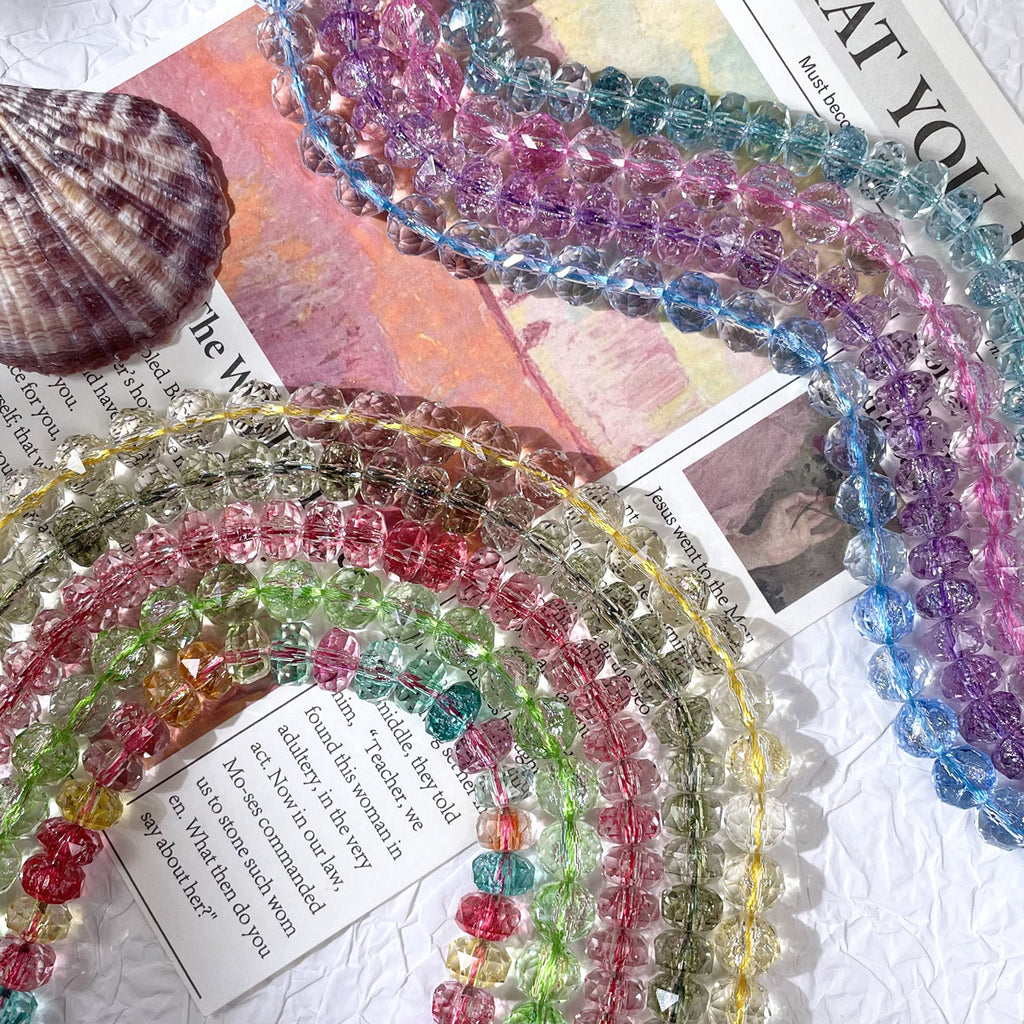 Strands of faceted crystal rondelle beads in assorted translucent colors on a white surface with a seashell and paper props
