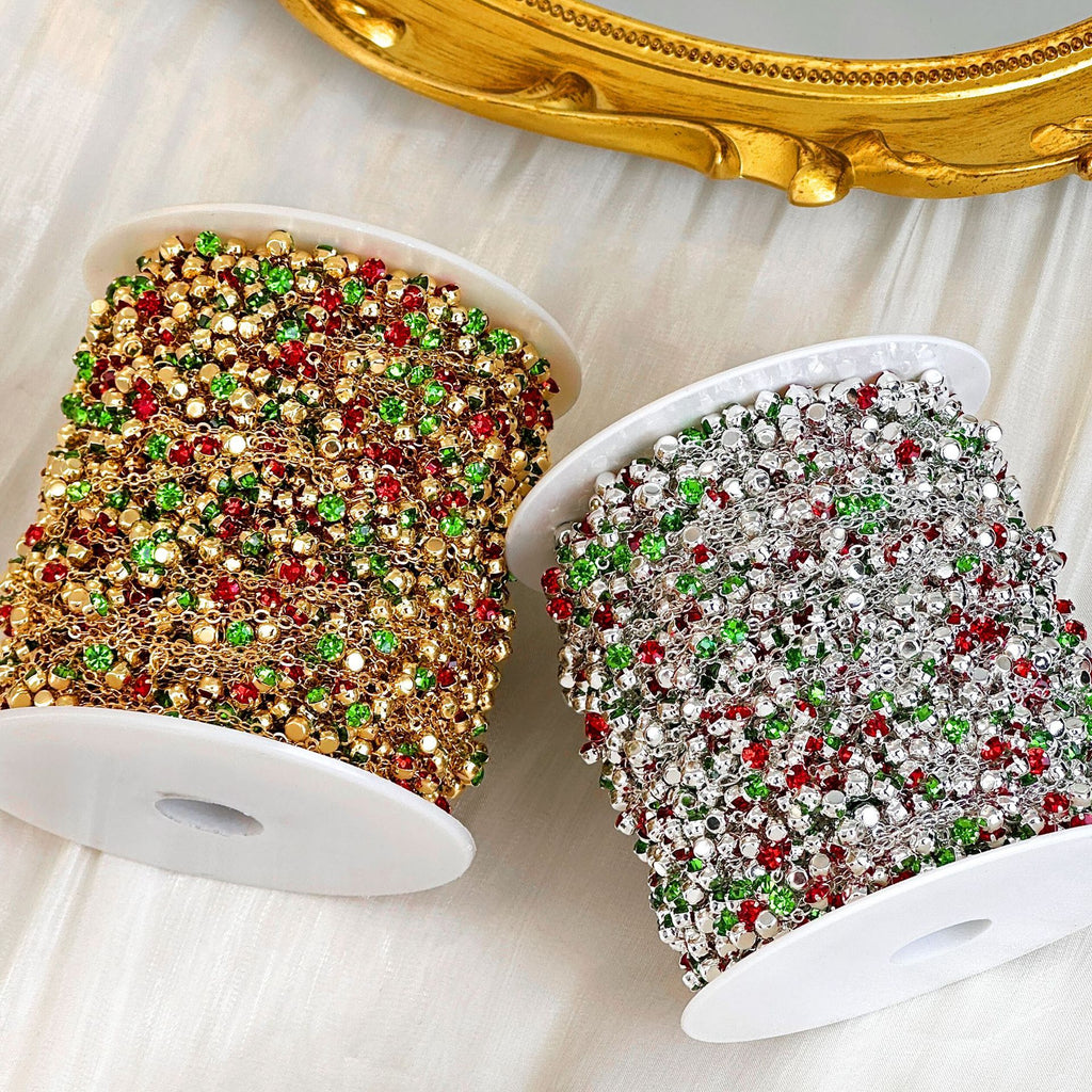 Two spools of decorative Christmas-themed rhinestone ribbon on a white fabric background, with a gold mirror frame partially visible at the top. One spool is gold with red and green accents, and the other is silver with red and green accents.