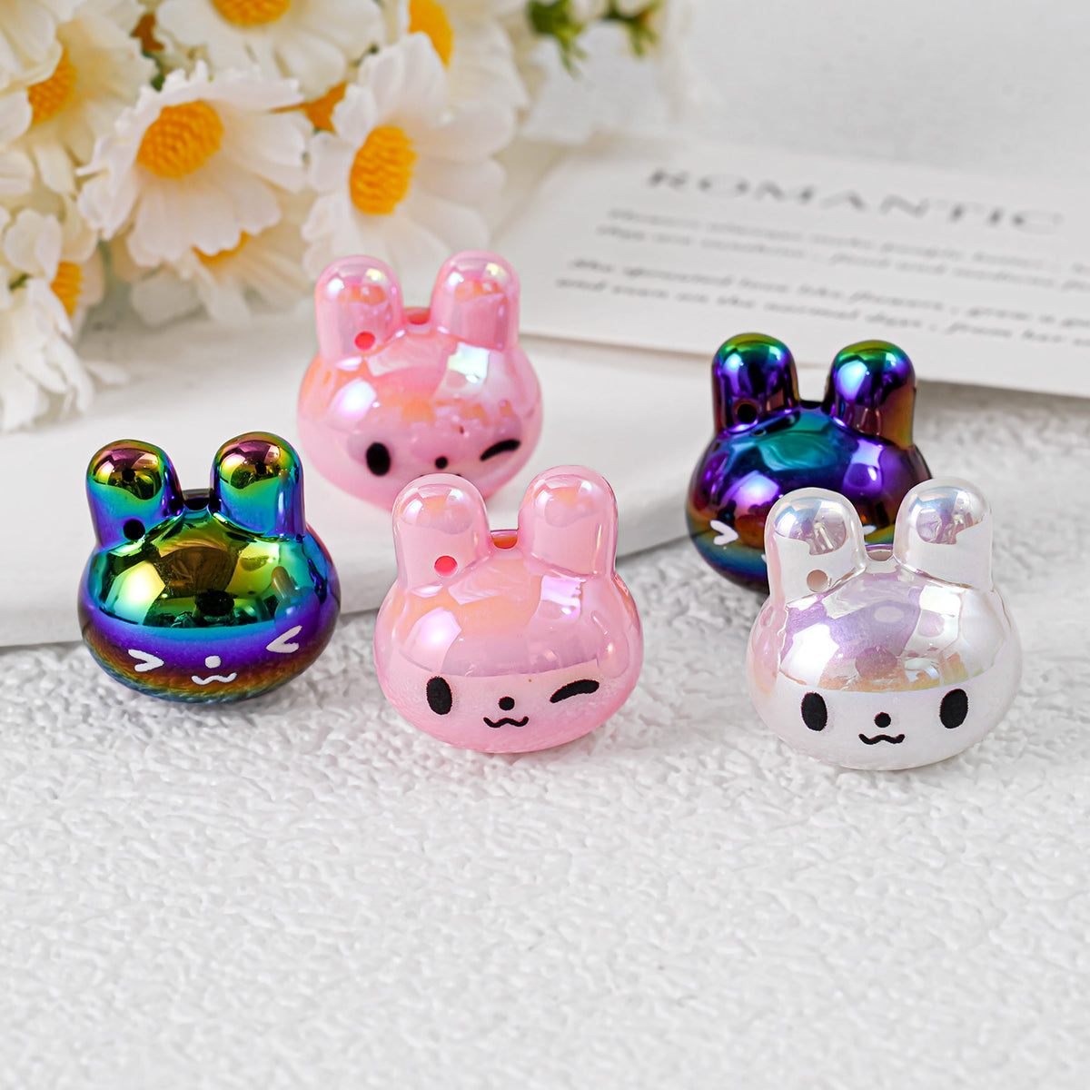 Five glossy bunny-shaped acrylic charms in rainbow, pink and silver-white on white surface with daisies