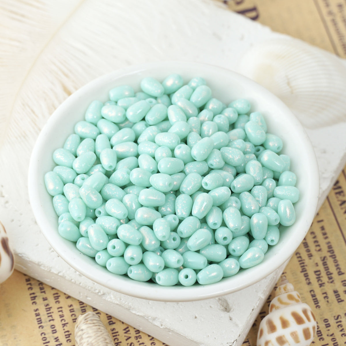 Light mint green glass teardrop beads in a white ceramic bowl on vintage newspaper with seashells