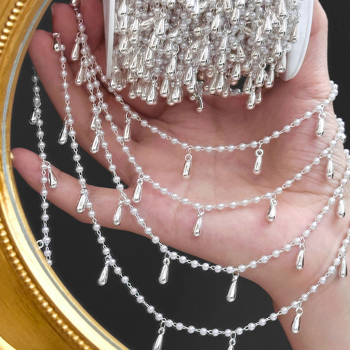 Hand holding spool of silver beaded fringe chains with teardrop drops against a dark background