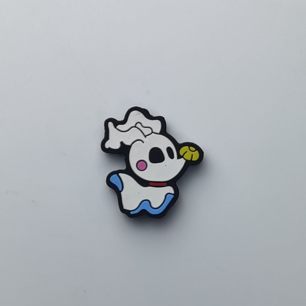 A small cartoon ghost-shaped accessory with a white body, black outline, blue lower edge, pink cheek, yellow mouth, and a white hat, photographed on a plain light gray background.