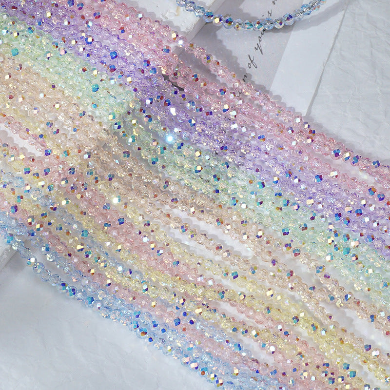 A close-up of iridescent rhinestone trim draped diagonally across a white textured surface. The trim features a pastel rainbow gradient of pink, purple, blue, yellow, and green crystals with reflective, multicolored sparkle.