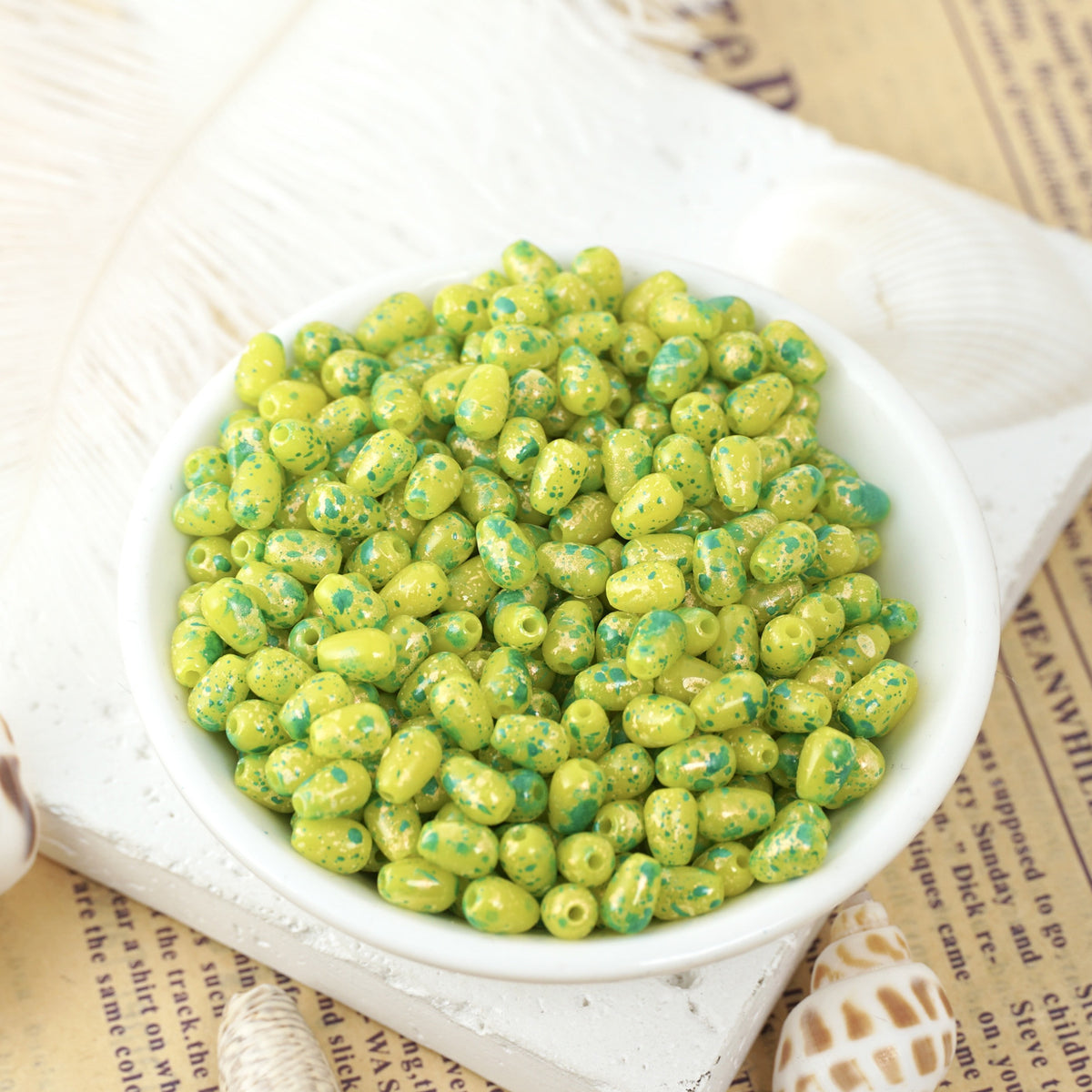 Green and yellow speckled glass teardrop beads in a small white bowl on vintage newspaper with seashells nearby