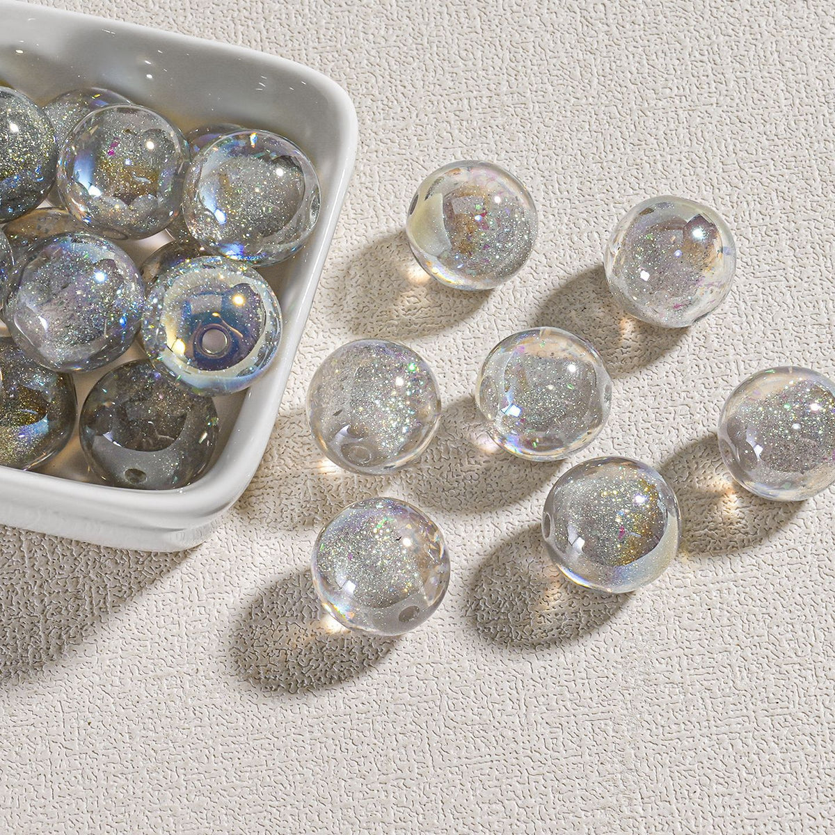 Clear round candy-core opal-effect resin beads with iridescent glitter scattered on textured beige surface