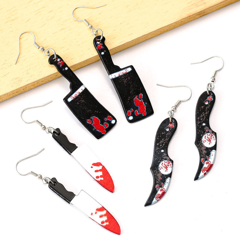 A set of gothic-style dangling earrings displayed on a white background with a light wood block. The earrings feature three designs: a pair of black cleaver-shaped earrings with red splatters and white text, a pair of silver-and-white knife-shaped earrings with red splatters, and a pair of black dagger-shaped earrings with red and white splatter details.
