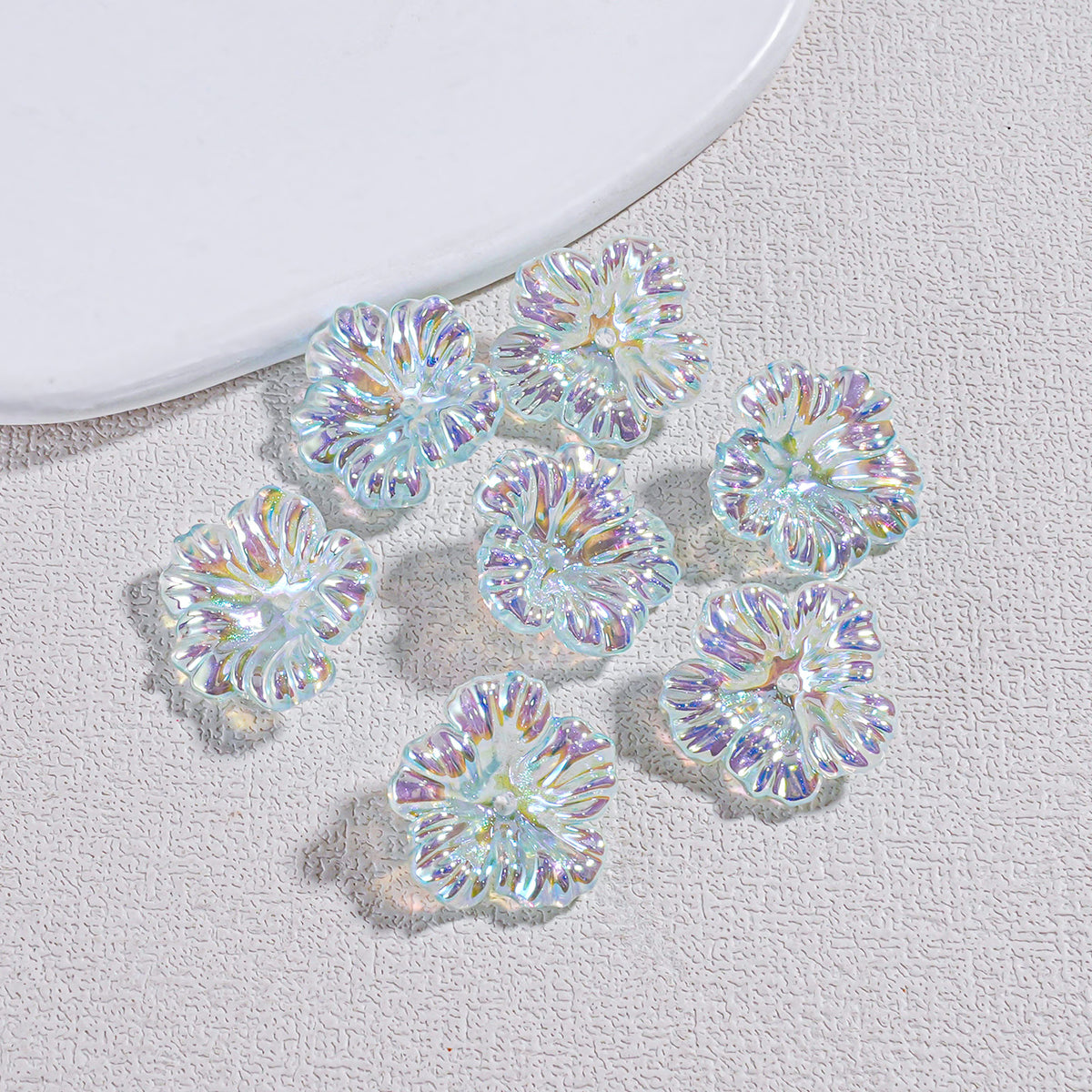 Seven iridescent holographic resin flower beads scattered on a textured gray surface by a white plate edge
