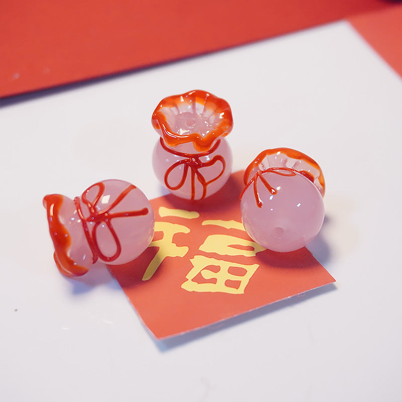 Three small translucent pink glass charms shaped like money bags with orange-red trim and bow details, arranged on a white surface with a red paper slip featuring a gold Chinese character.