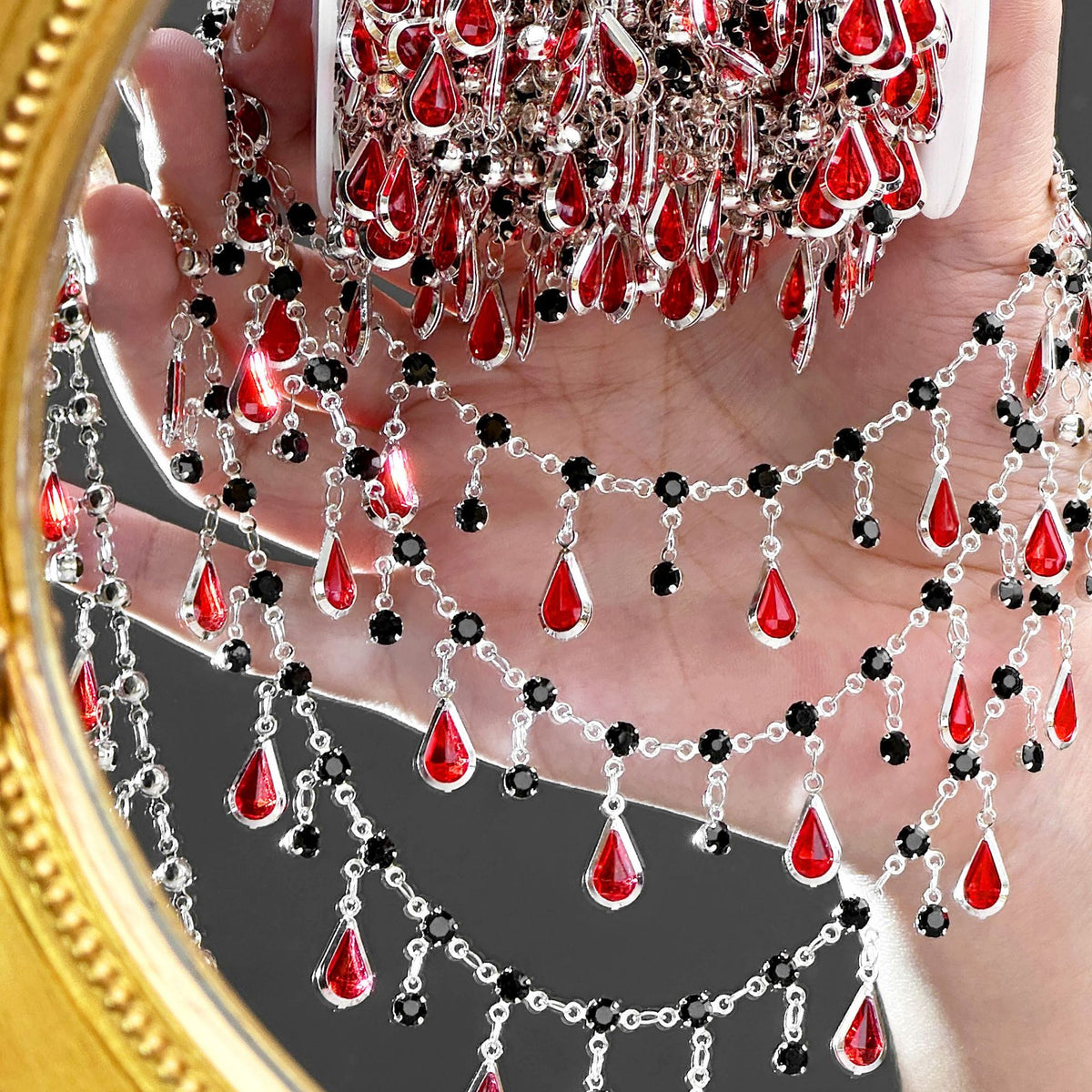 A hand holds a large silver-toned jewelry stand covered with multiple layered silver chains draped with red teardrop-shaped crystals and small black round stones. A gold-framed mirror edge is visible on the left, reflecting the jewelry setup.