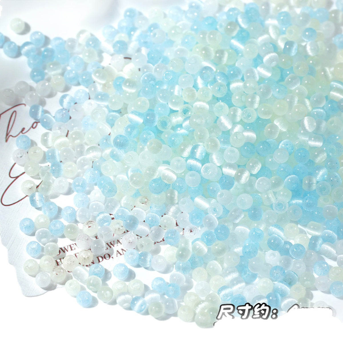 A pile of small round translucent beads in pale aqua blue, white, and milky clear, scattered on a white surface with a decorative printed napkin partially visible underneath.