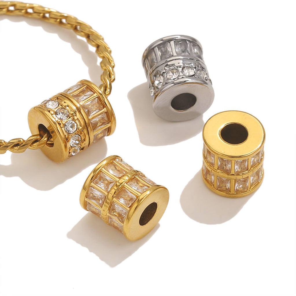 Four cylindrical charm beads with clear crystal accents, shown in gold and silver finishes on a white background. One gold bead is threaded onto a twisted gold chain.