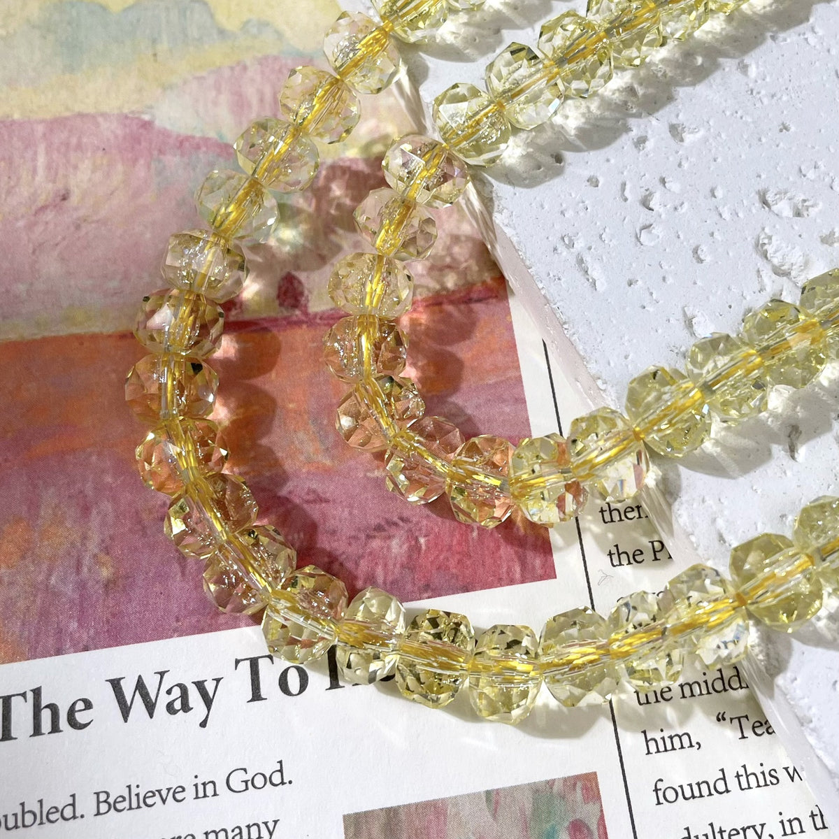 Strands of faceted clear beads with a soft yellow tint on a gold wire, on a textured white surface and printed paper