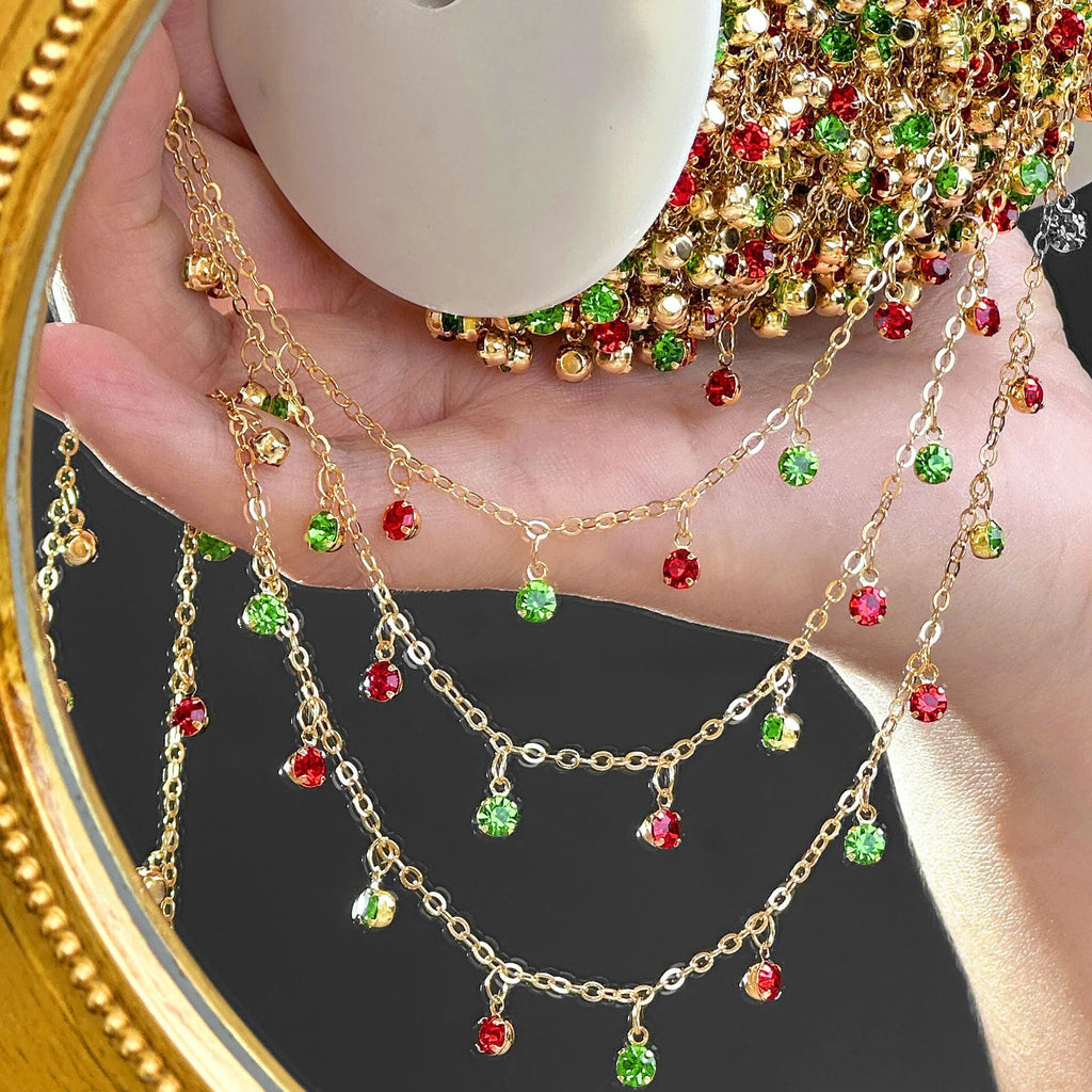 A gold-tone double-layered chain necklace with dangling rhinestone charms in red, green, and clear, held in a hand in front of a mirror.