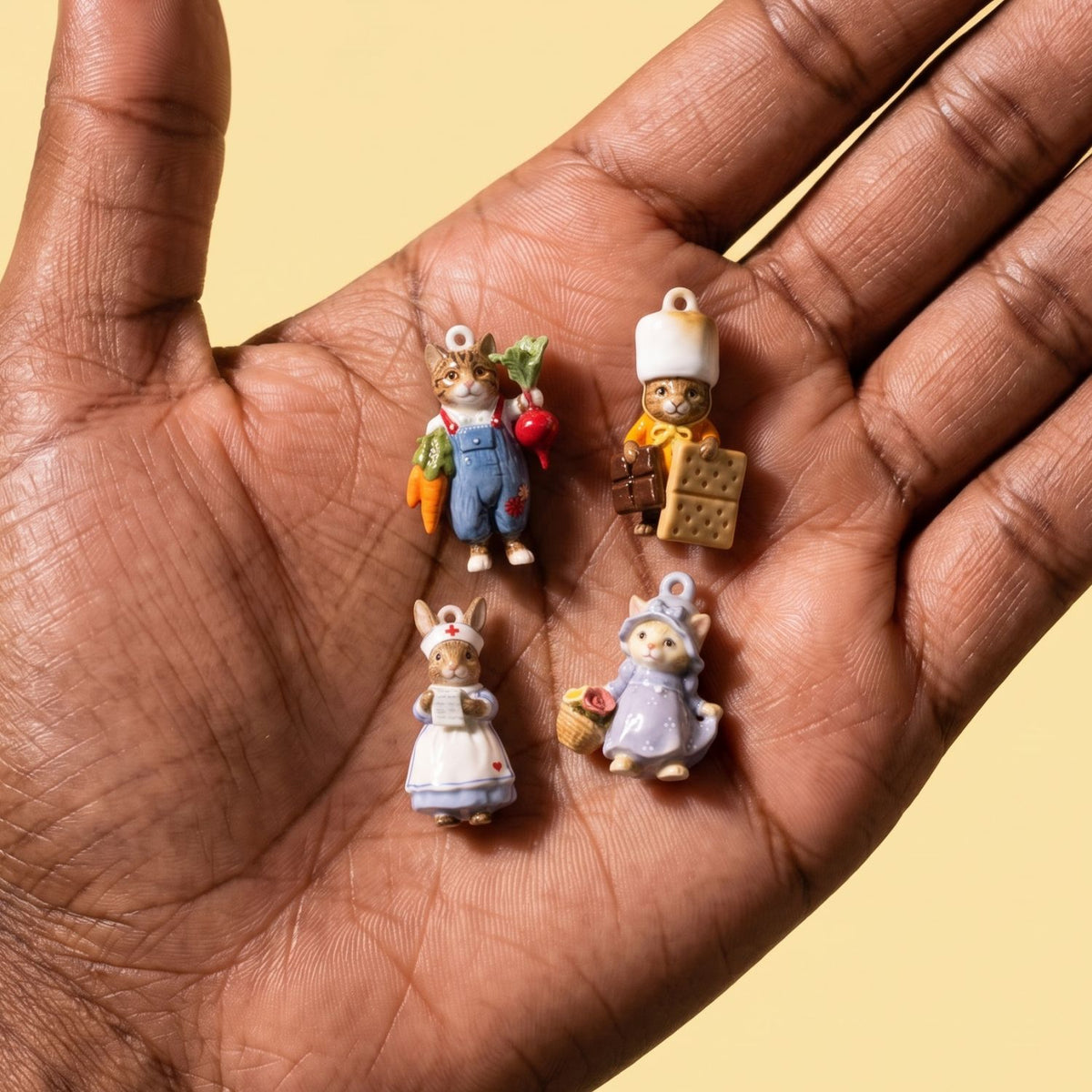 Hand holding four 3D animal charms: farmer cat, chef with biscuits, nurse rabbit, and cat in purple dress with basket.