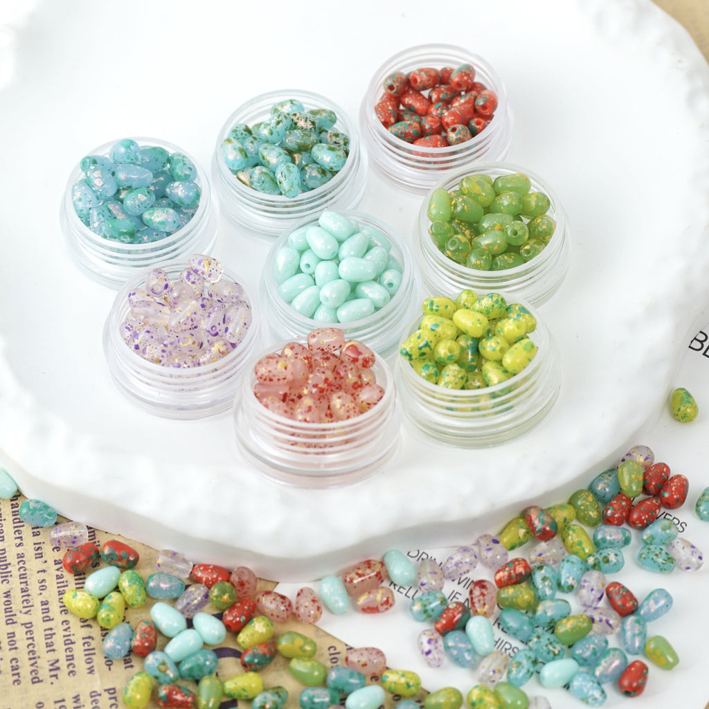 Clear jars of colorful speckled glass teardrop beads on a white scalloped plate with loose beads scattered
