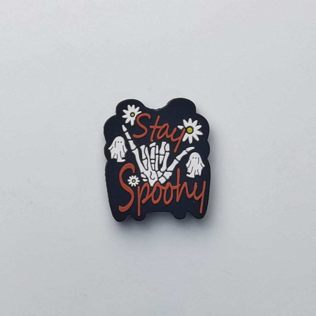 A black Halloween-themed enamel pin featuring a skeleton hand holding daisies, white ghosts, and the words 'Stay Spooky' in orange script, shown on a plain light gray background.