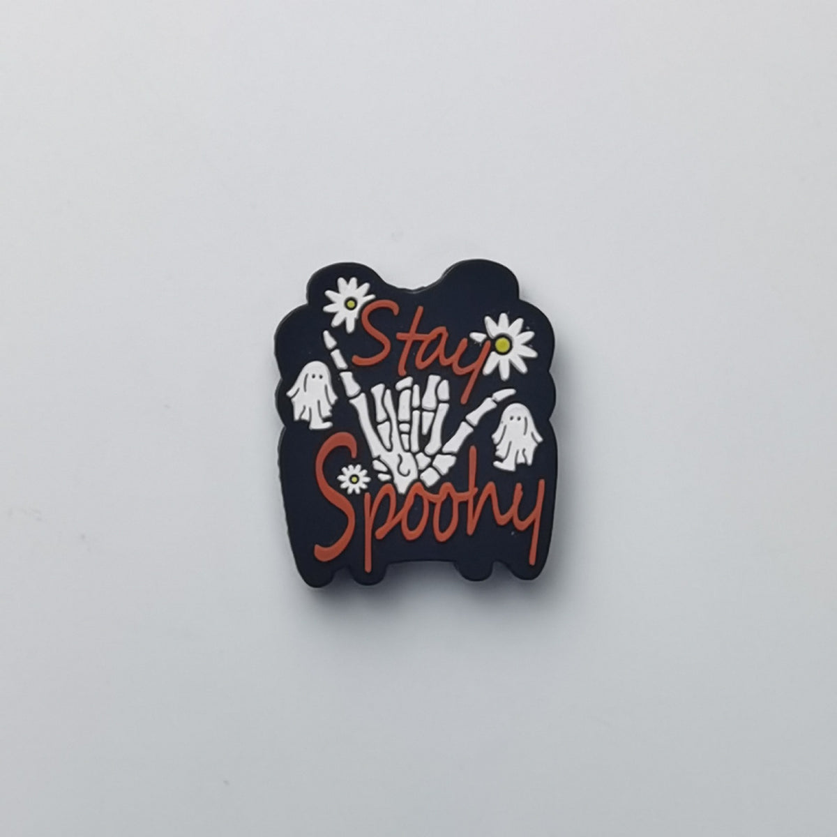 A black Halloween-themed enamel pin featuring a skeleton hand holding daisies, white ghosts, and the words 'Stay Spooky' in orange script, shown on a plain light gray background.